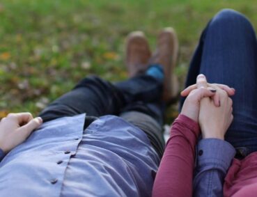A romantic couple holds hands while lying on grass during a sunny day in the park.